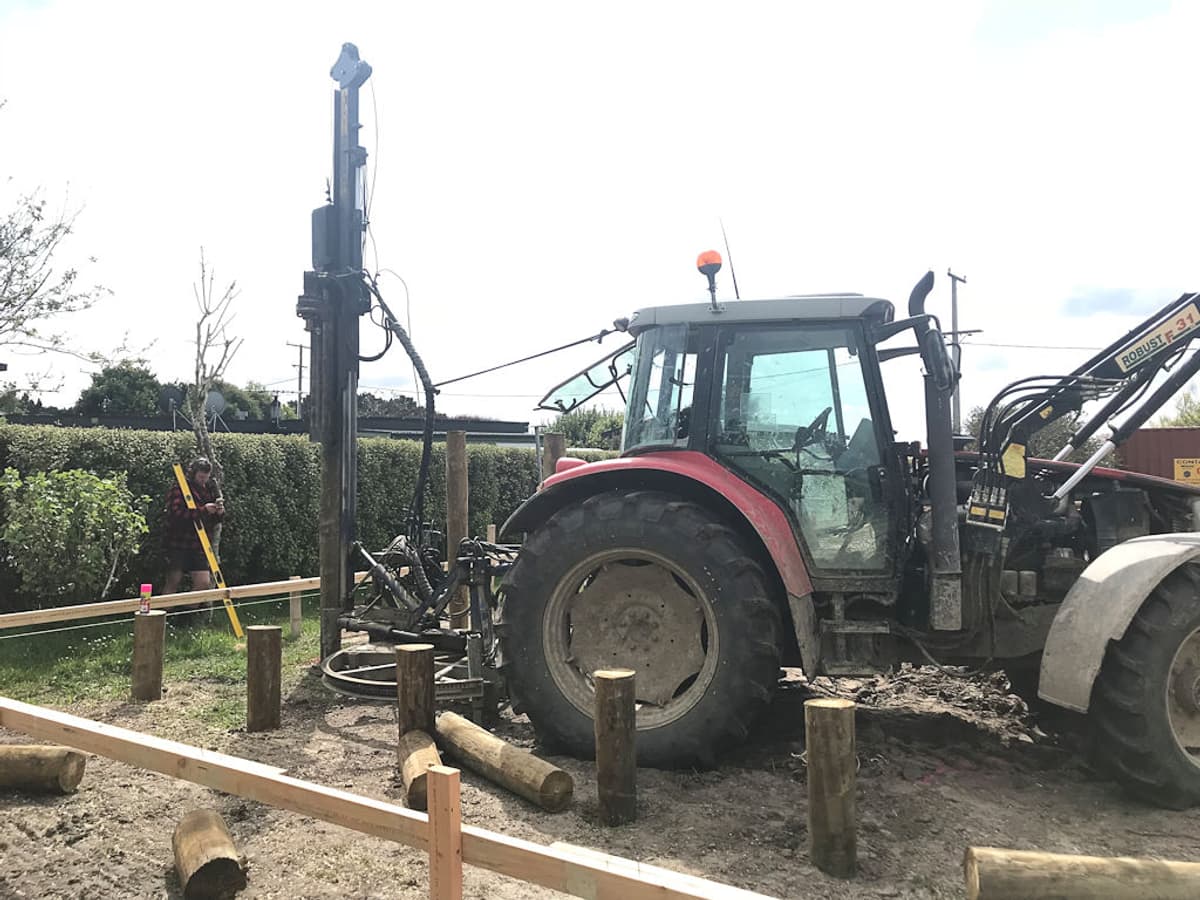 Tractor-mounted hydraulic driver setting timber piles to a string line on a levelled residential site, with a worker holding a staff at the far peg