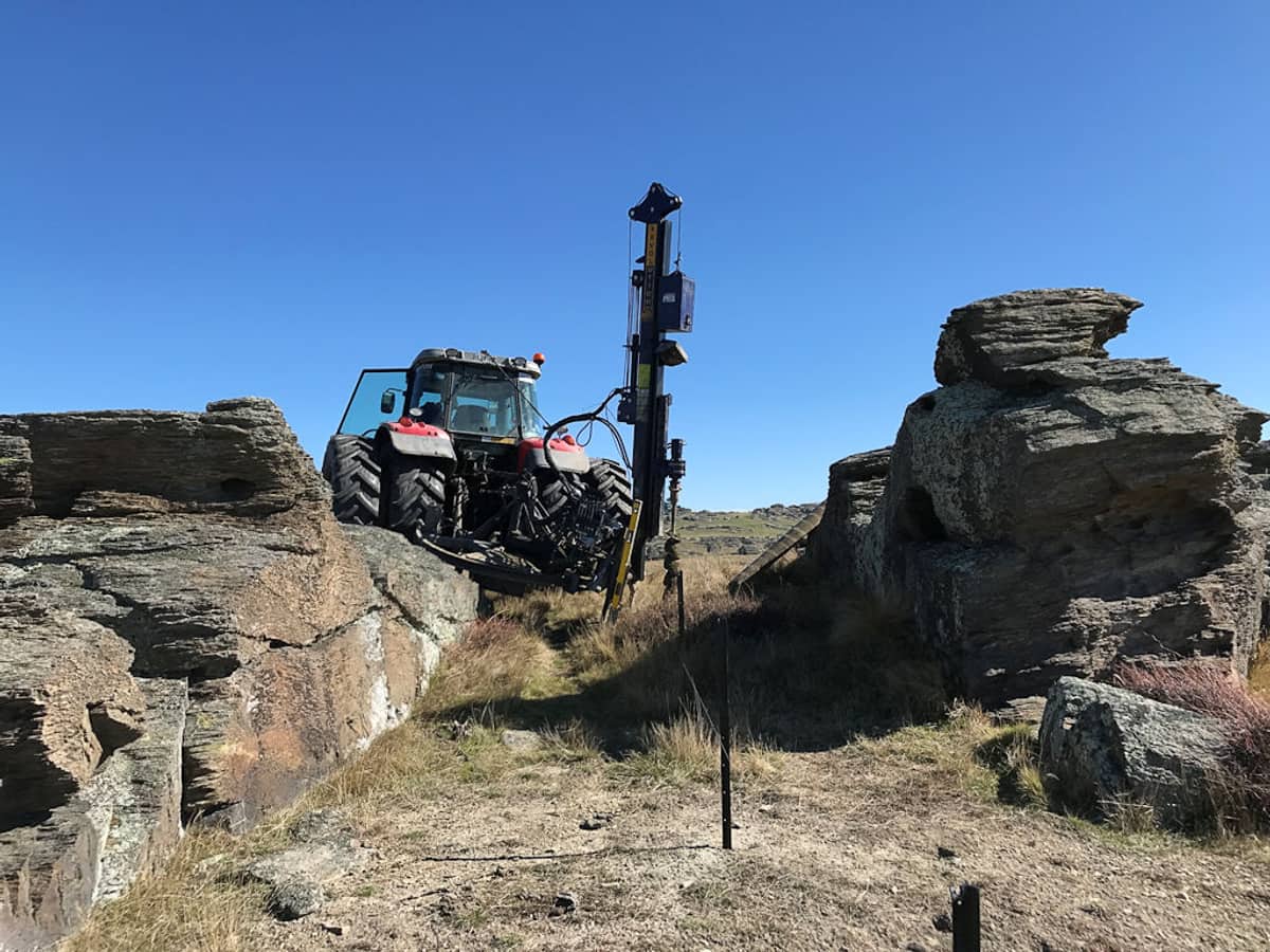 Tractor-mounted post driver working between weathered schist tors on a dry hillside, driving a line of steel standards through rock